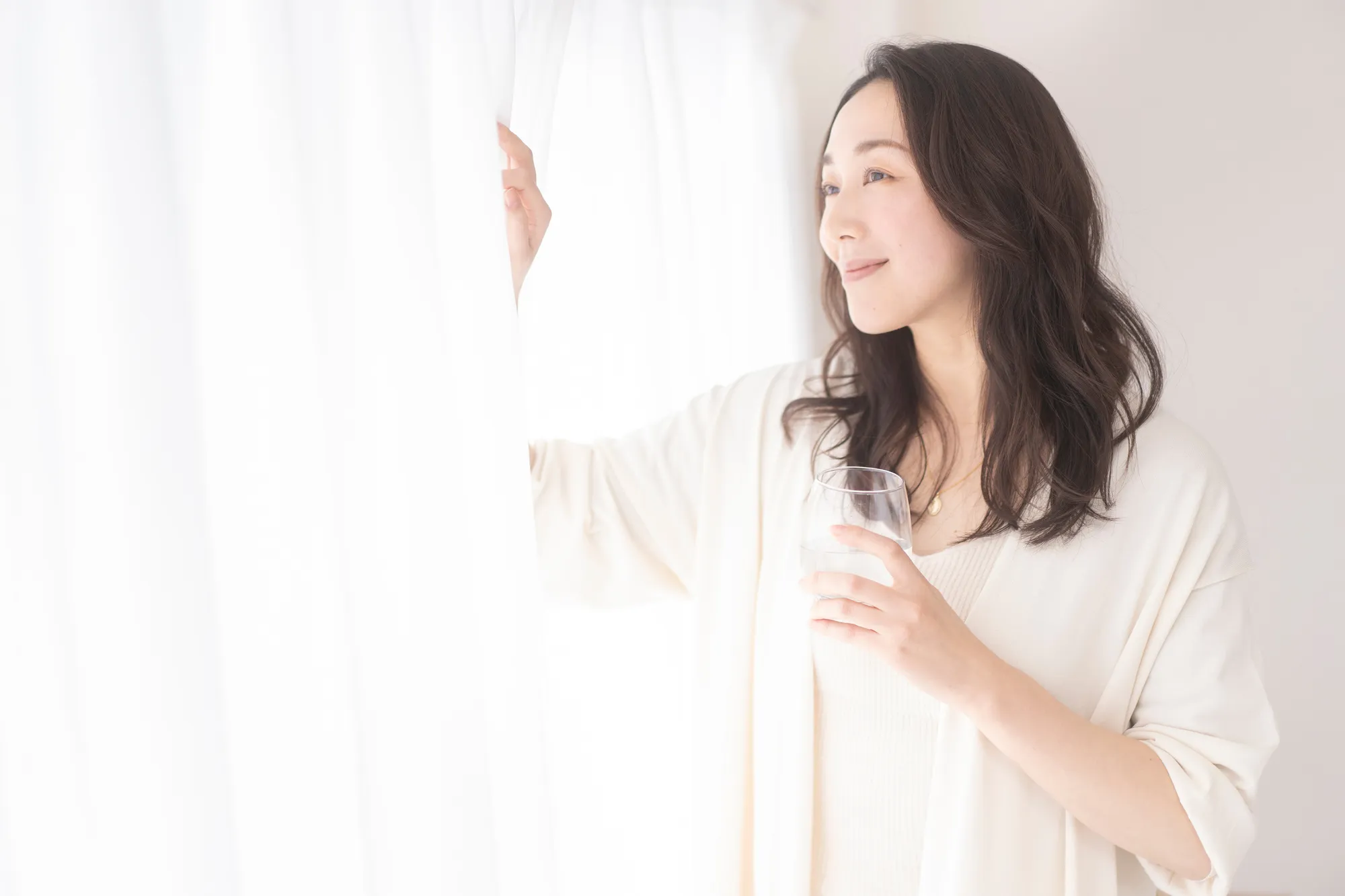 朝カーテンをあけて水を飲む女性